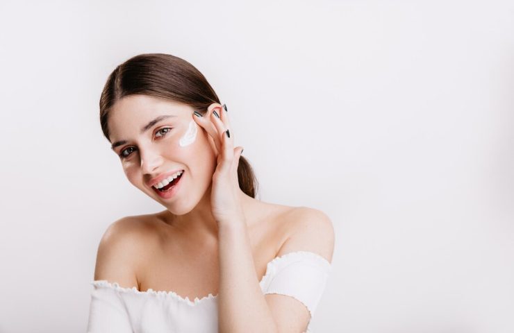 smiling-green-eyed-girl-puts-cream-clean-face-brunette-white-top-posing-isolated