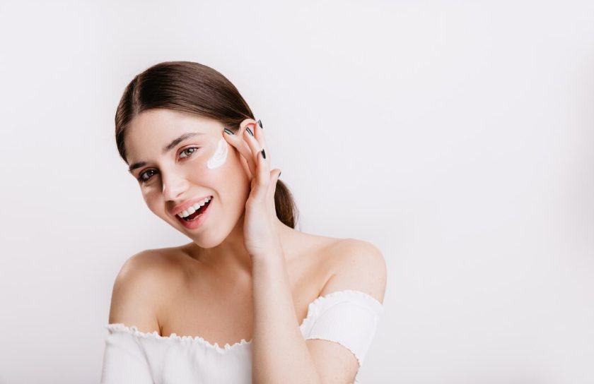 smiling-green-eyed-girl-puts-cream-clean-face-brunette-white-top-posing-isolated